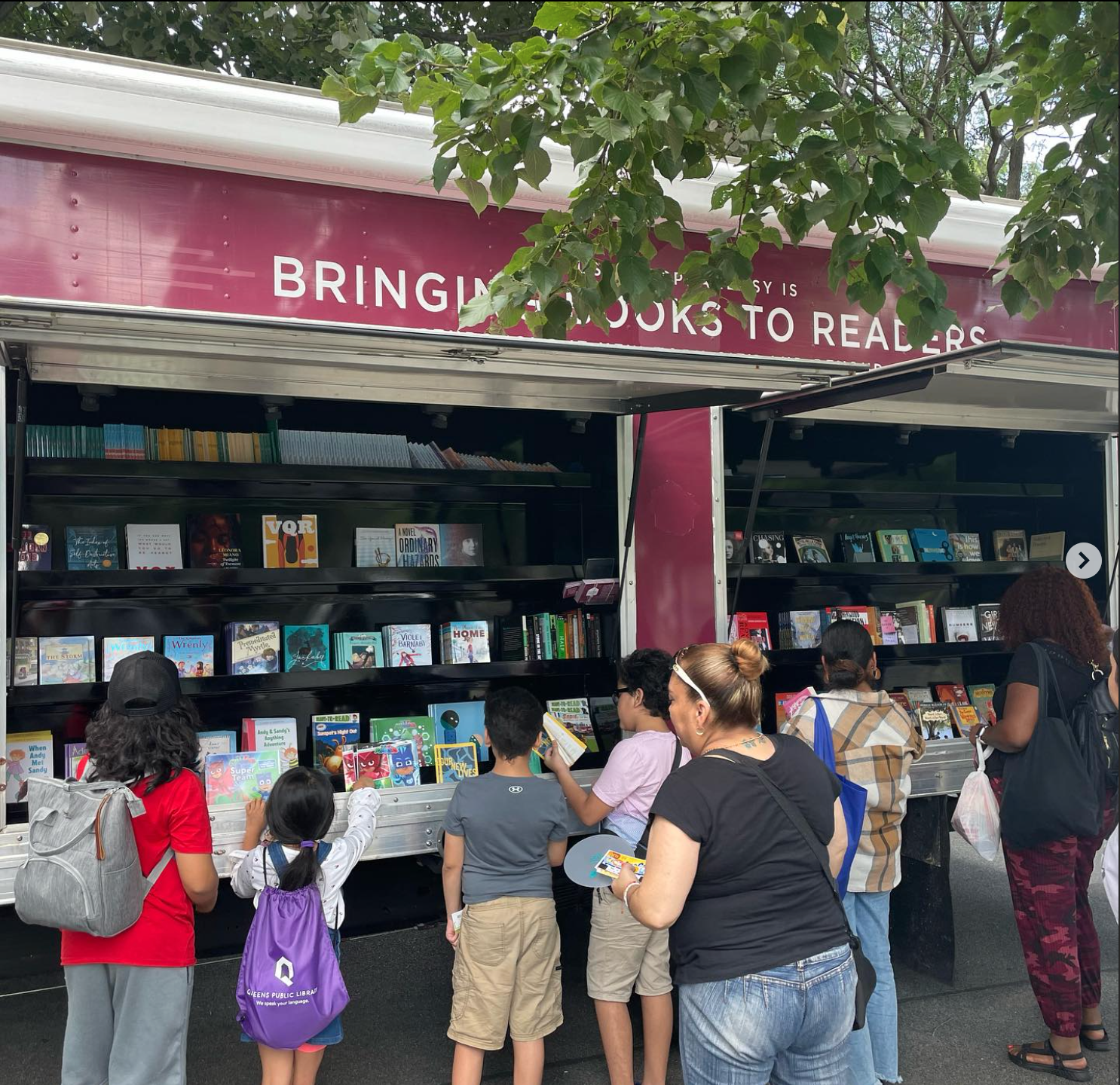 House of SpeakEasy's Bookmobile, parked on a sunny street behind a rack of bikes. A crowd of people is gathered by the Bookmobile's shelves, choosing their books.