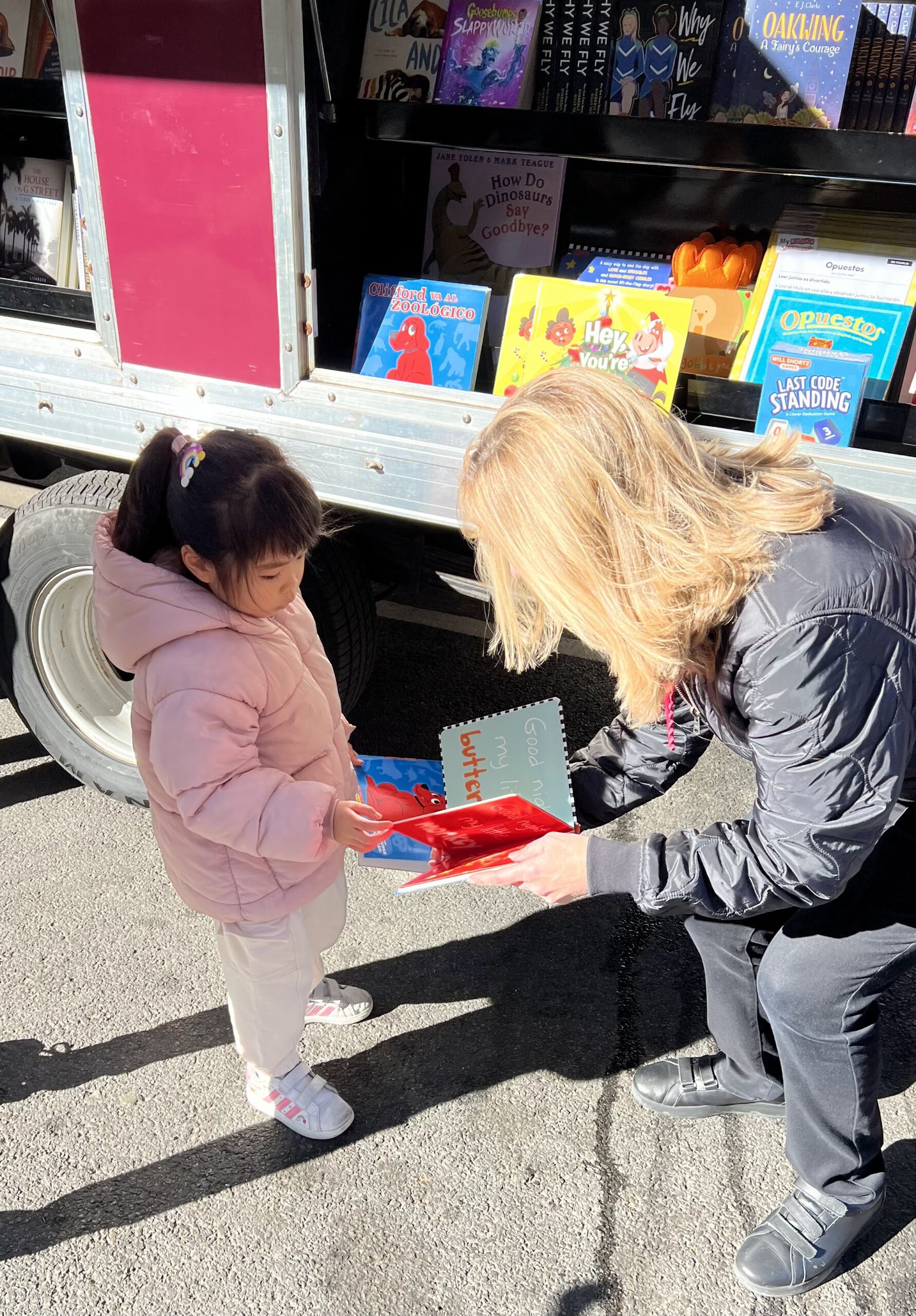 House of SpeakEasy's Bookmobile, parked on a sunny street behind a rack of bikes. A crowd of people is gathered by the Bookmobile's shelves, choosing their books.