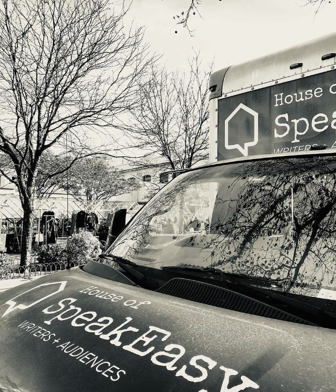 House of SpeakEasy's Bookmobile, parked on a sunny street behind a rack of bikes. A crowd of people is gathered by the Bookmobile's shelves, choosing their books.