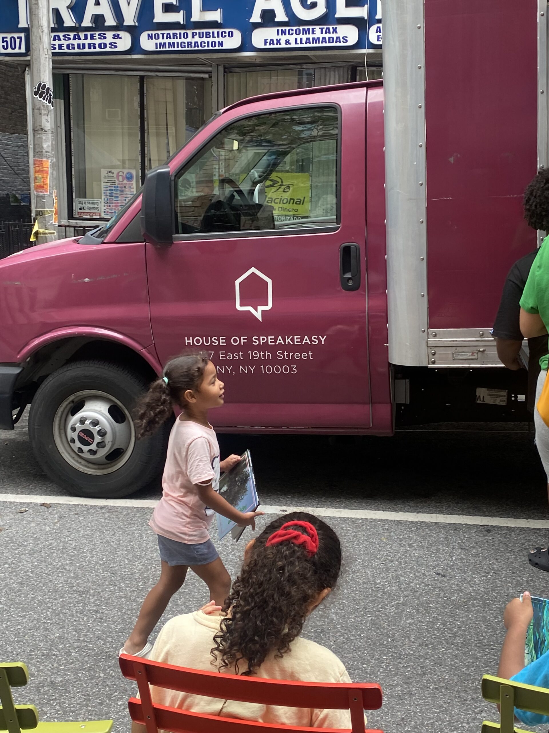 House of SpeakEasy's Bookmobile, parked on a sunny street behind a rack of bikes. A crowd of people is gathered by the Bookmobile's shelves, choosing their books.