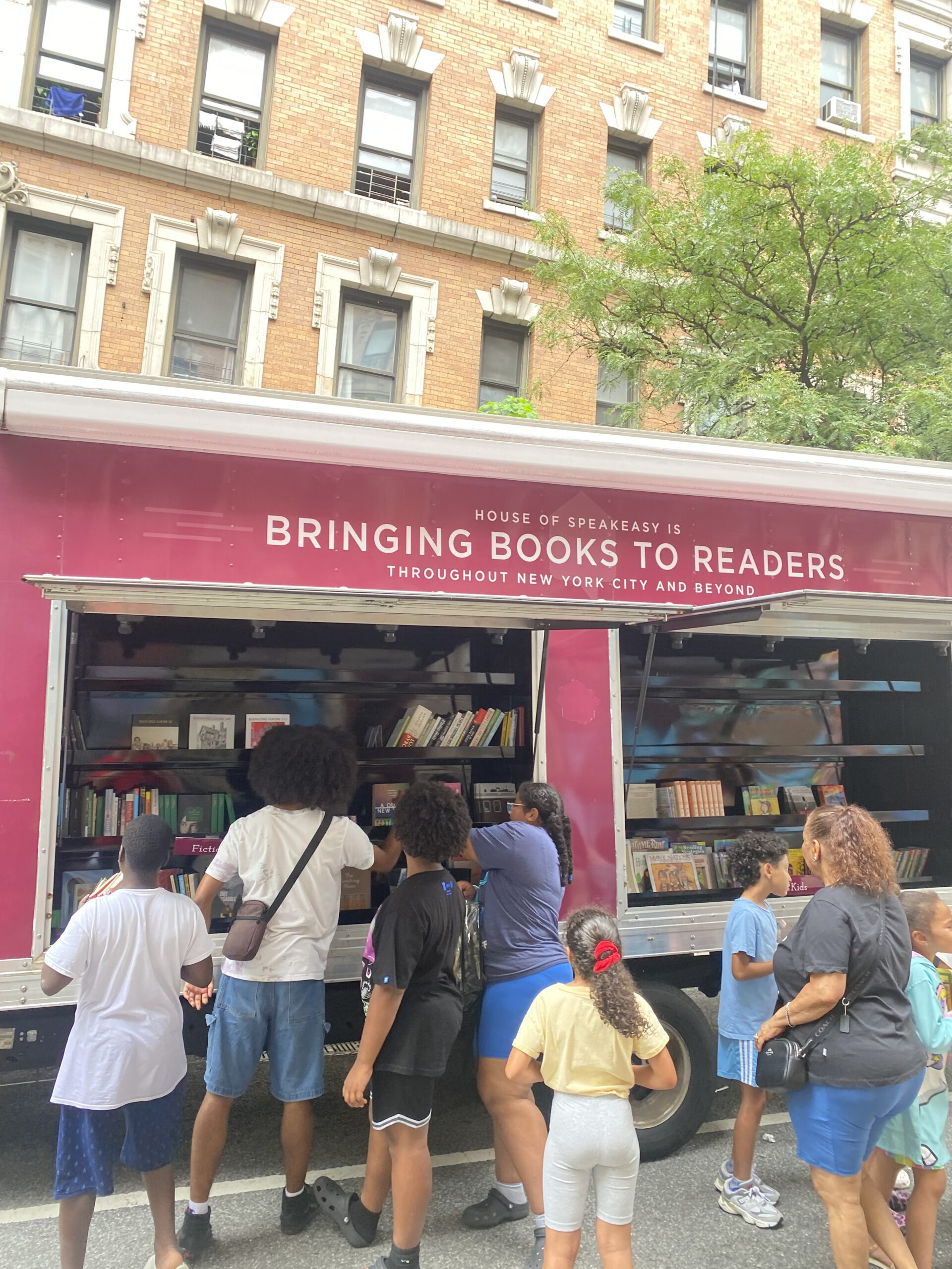 House of SpeakEasy's Bookmobile, parked on a sunny street behind a rack of bikes. A crowd of people is gathered by the Bookmobile's shelves, choosing their books.