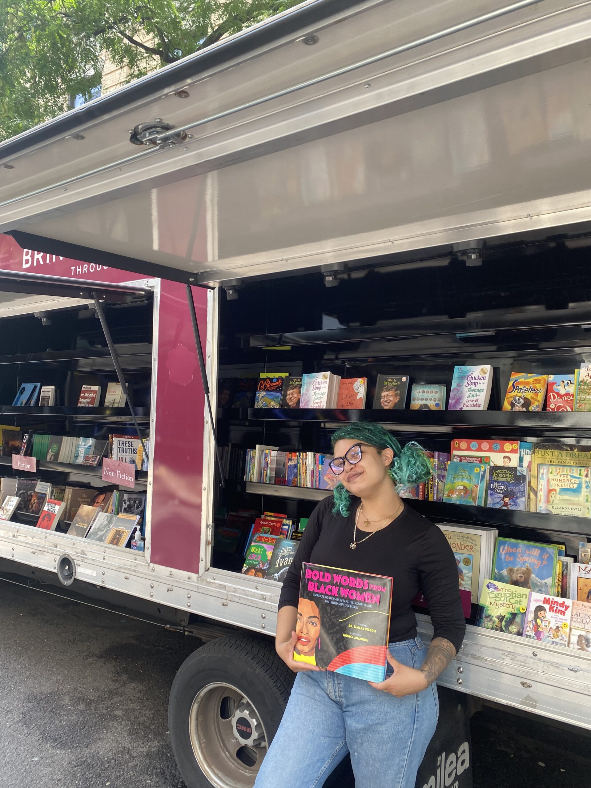 House of SpeakEasy's Bookmobile, parked on a sunny street behind a rack of bikes. A crowd of people is gathered by the Bookmobile's shelves, choosing their books.