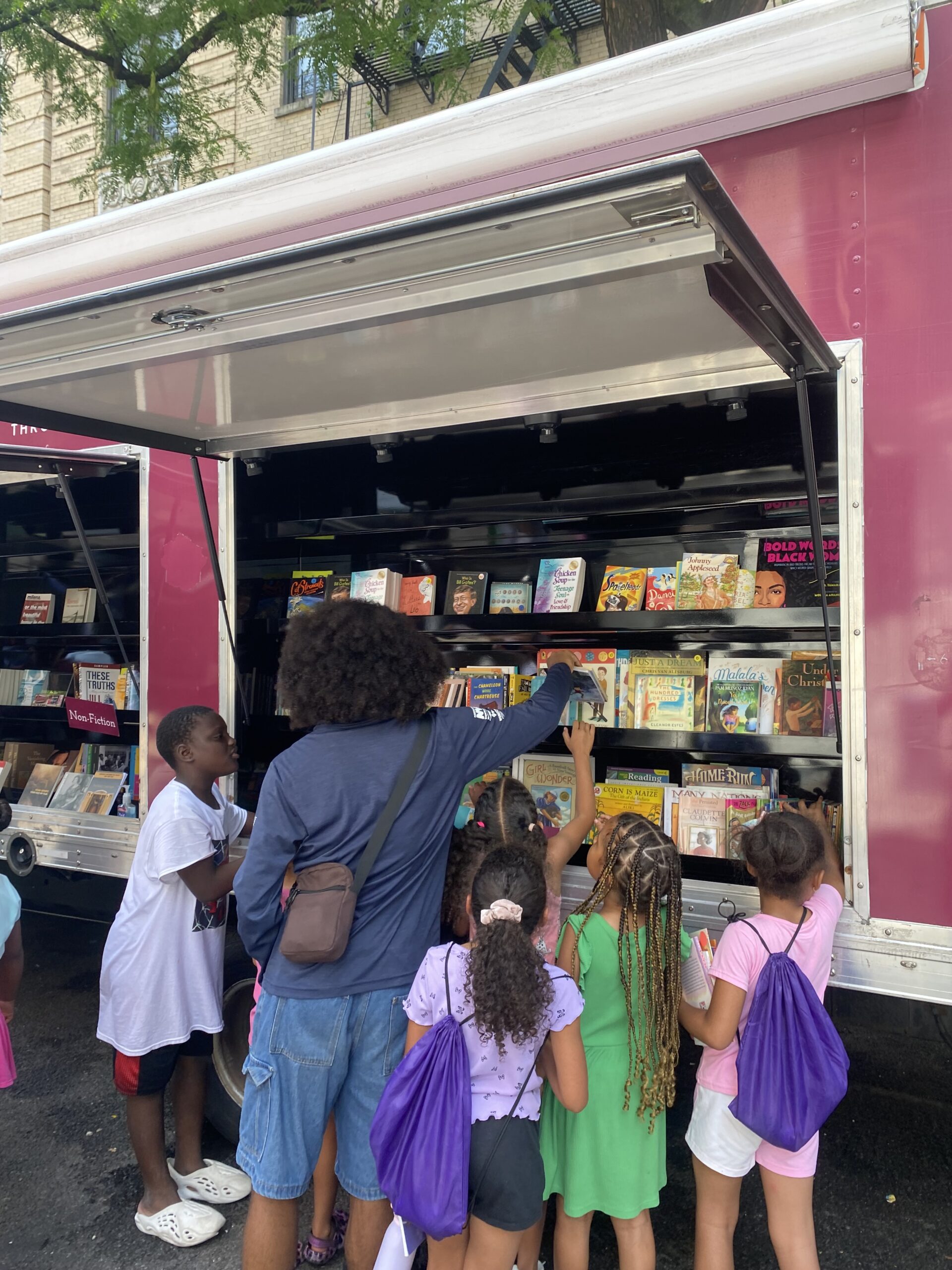 House of SpeakEasy's Bookmobile, parked on a sunny street behind a rack of bikes. A crowd of people is gathered by the Bookmobile's shelves, choosing their books.